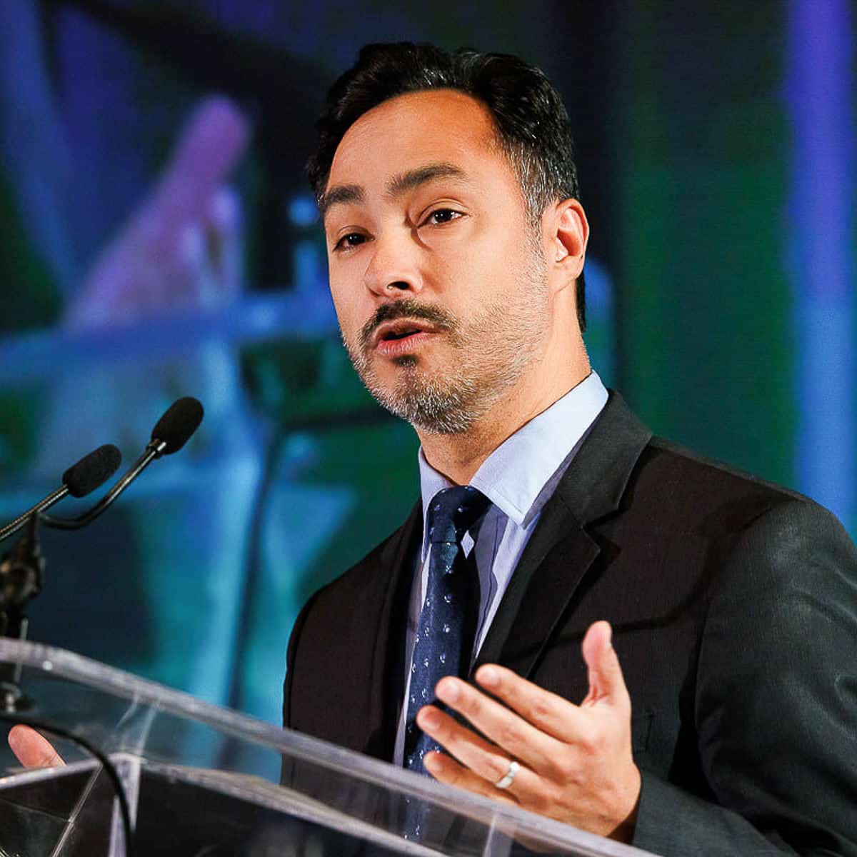 A keynote speaker in a suit stands at the podium, confidently addressing the audience through two microphones.