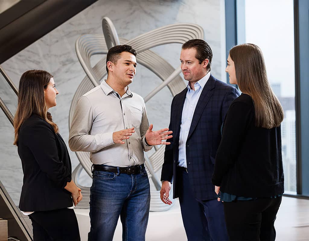 Four people standing and talking in a modern office with a large abstract sculpture in the background.
