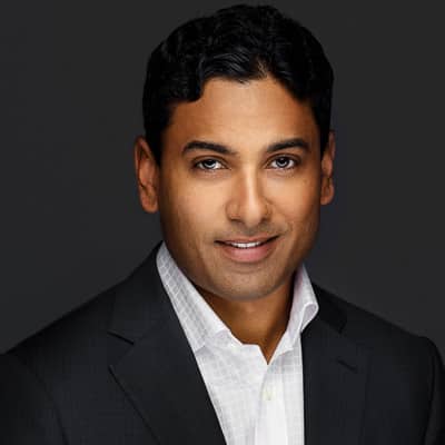 A corporate headshot of a man in a suit and checkered shirt smiling at the camera against a gray background.