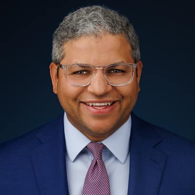 A corporate headshot of a smiling man with glasses wearing a blue suit and a patterned tie.